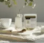 White cup, saucer, and vase with leafy branches on a table. A spoon and packet of Organic Tulsi Powder nearby. Soft lighting, calm mood. One of the herbs for Kapha.