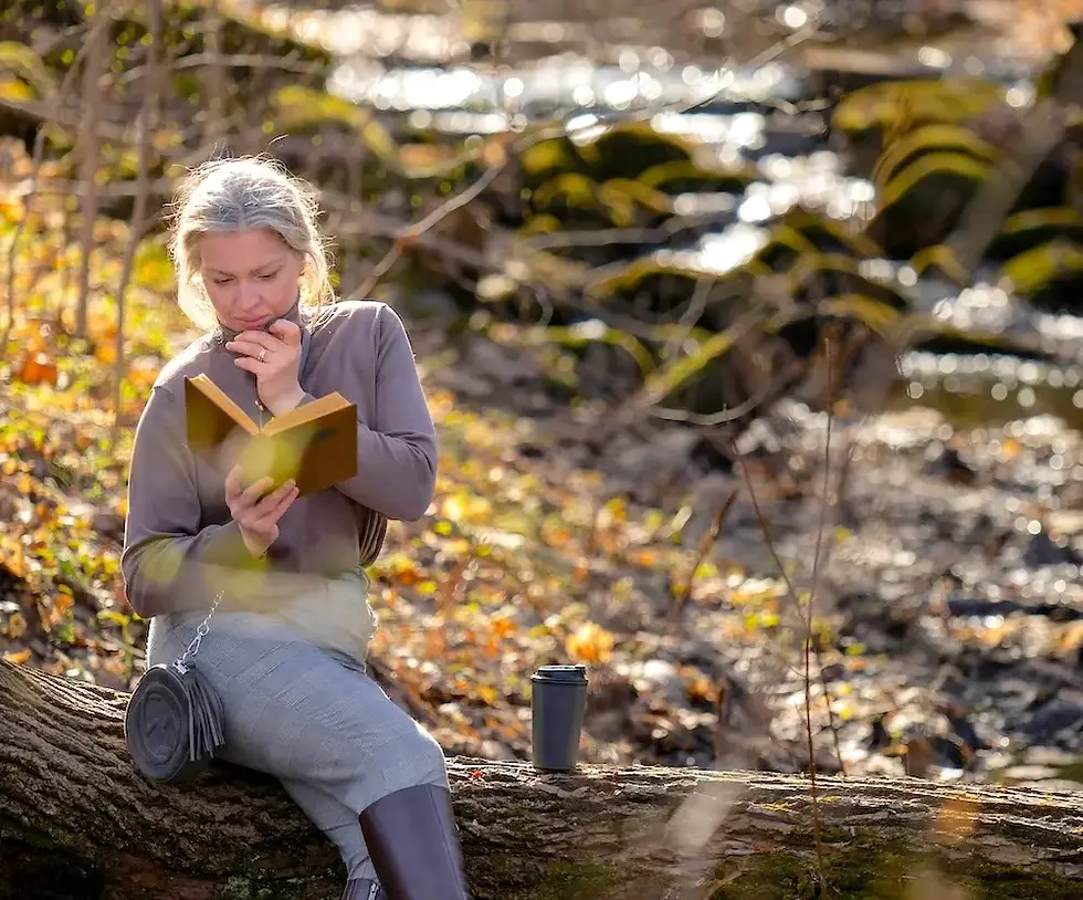 A woman over 40 sits on a log in a forest, reading a book thoughtfully as she contemplates the Ayurveda of fall. A travel mug is beside her. The background shows a sunlit woodland area during the Vata season Ayurveda.