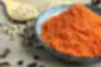 Blue bowl of vibrant red-orange spice on wooden table; scattered white and black peppercorns; wooden spoon. Rustic kitchen vibe for these Kapha pacifying foods.