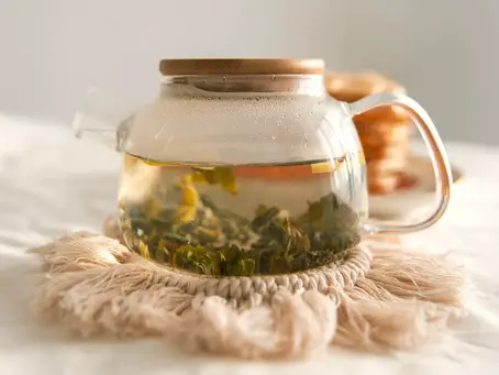 Pitta tea: Glass teapot with green herbs and hot water sits on a fringed mat. Soft, warm lighting creates a calm atmosphere.