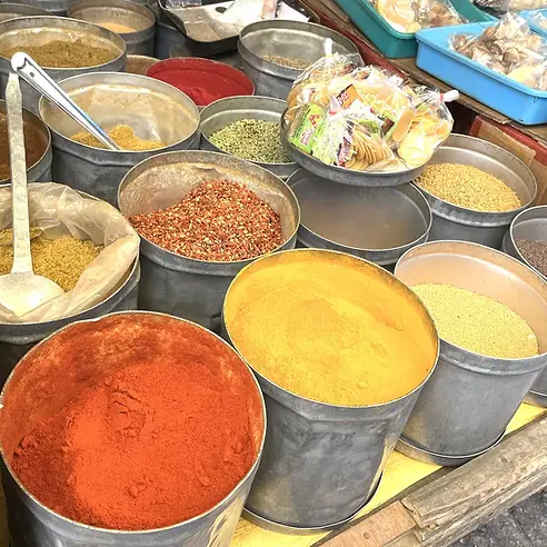 A vibrant display of traditional Indian spices overflowing from large buckets at an open-air stall in the Kalimpong Haat Market, West Bengal, India.