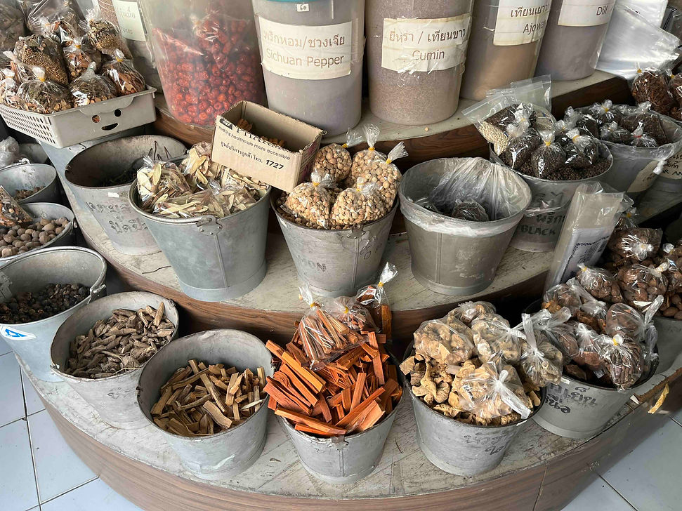 A vibrant outdoor herb and spice market stall in Chiang Rai, Thailand, displaying large silver buckets filled with traditional dried Ayurvedic herbs, cinnamon sticks, Sichuan pepper, and ground cooking spices.