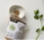 A bag of organic coriander seeds is beside a white bowl with some seeds, and fresh green cilantro leaves on a white background.