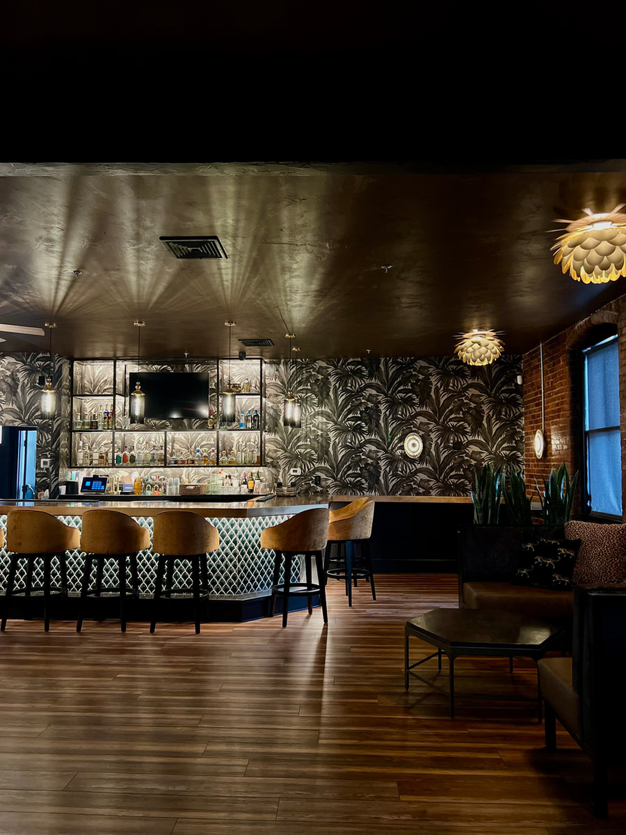 A dimly lit upscale bar featuring golden velvet barstools, an illuminated liquor display, and tropical pattern wallpaper.