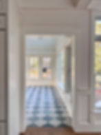 An doorway view looking into a bright sunroom featuring a grey and white checkerboard tile floor, large paned windows, and white wall trim.