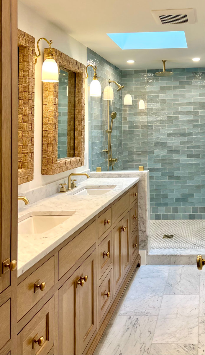 A luxury bathroom featuring a long light wood double vanity with marble countertops, framed mirrors, and a walk-in shower with light blue subway tiles and a skylight.