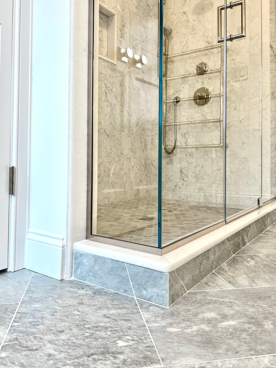 A luxury walk-in glass shower featuring white marble wall tiles, a built-in shelf, brushed metal fixtures, and matching grey marble floor tiles.