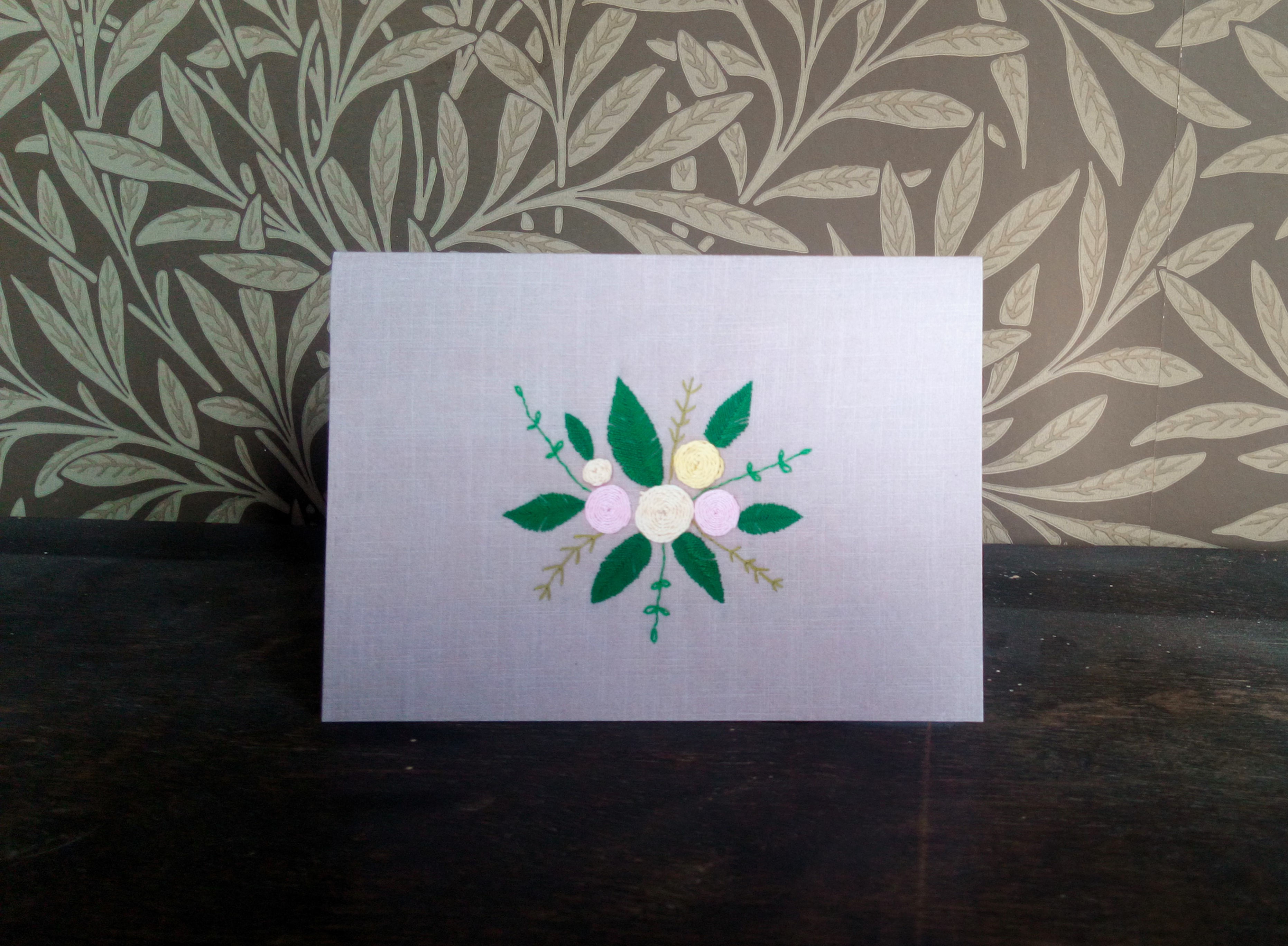Greetings card featuring hand embroidered textile artwork of a bloom of flowers and leave on a dark ecru fabric background