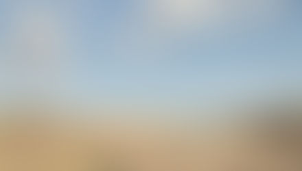 Grassy path leads to a river under a bright blue sky with fluffy clouds. Trees and dry reeds surround, creating a serene atmosphere.