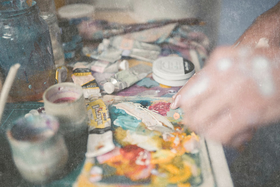 A hand mixes paint on a colorful palette with scattered paint tubes in a messy art studio. Jars and brushes are visible in the background.
