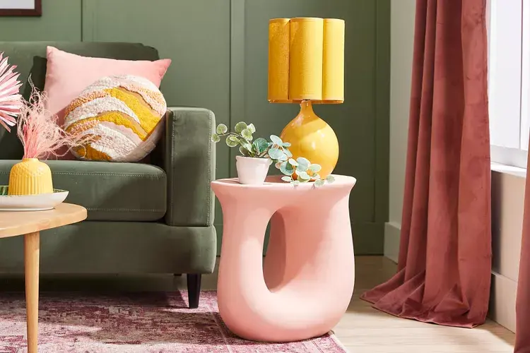 Светла и модерна дневна стая, обзаведена със зелено канапе и декоративни възглавници в пастелни цветове. Включва уникална розова масичка с модерна жълта лампа и малко растение, допълнени с розови завеси и мек килим за уютна атмосфера.