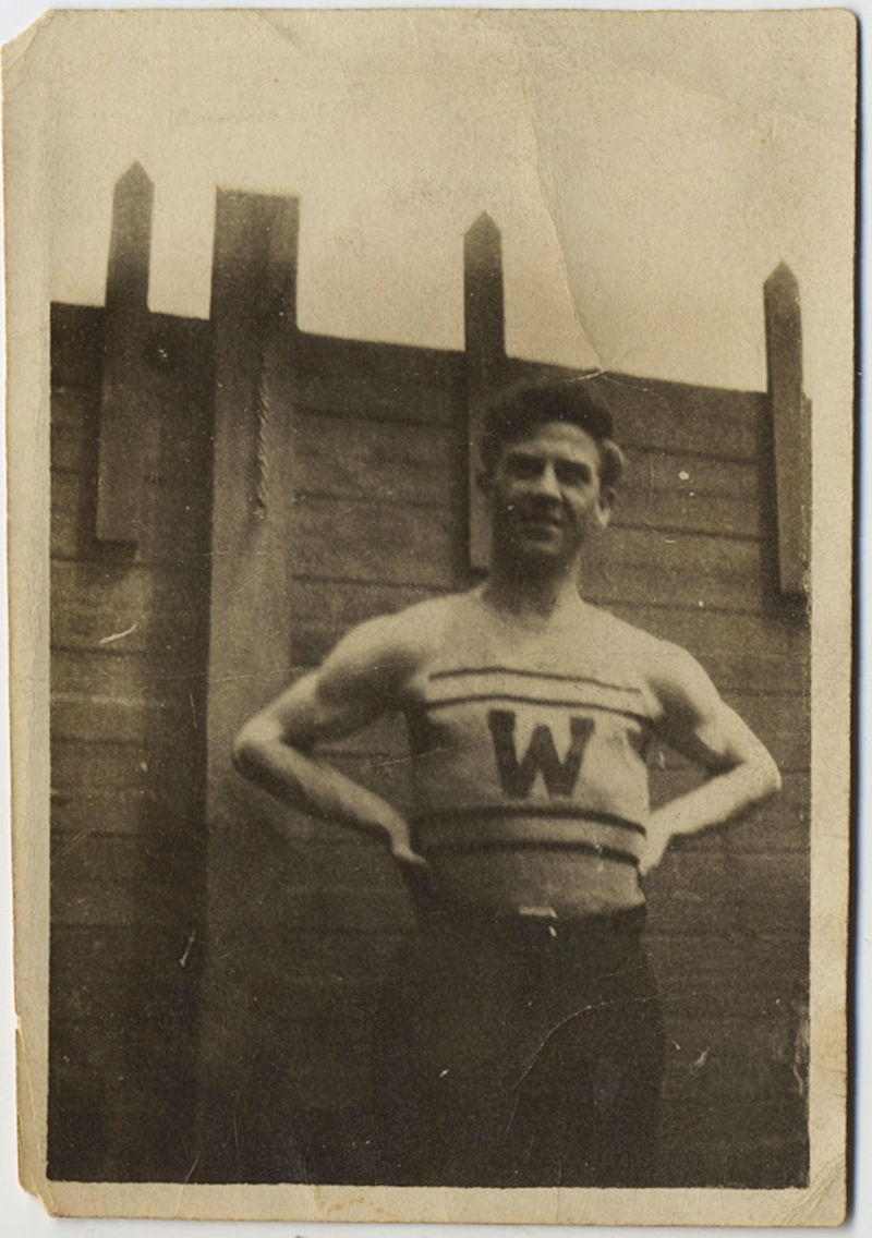 ATHLETE in LETTER VEST "W" against WOODEN FENCE ARMS AKIMBO