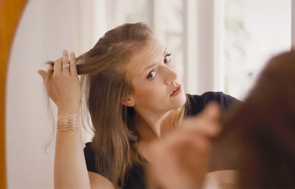 Woman struggling with hair loss in front of her hair and looking at the damage in the mirror, wearing silver bracelets. Soft lighting in a neutral-toned room. Focused expression.