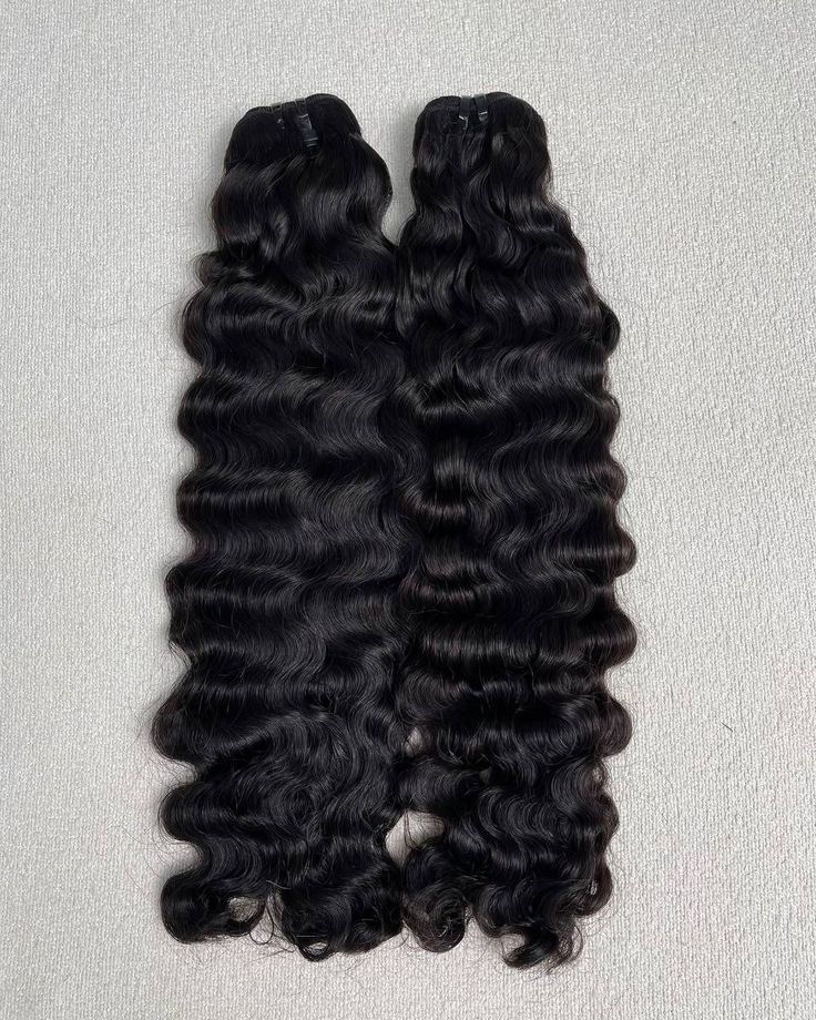 Two bundles of glossy black wavy hair extensions on a light gray surface, showcasing natural waves and shine.