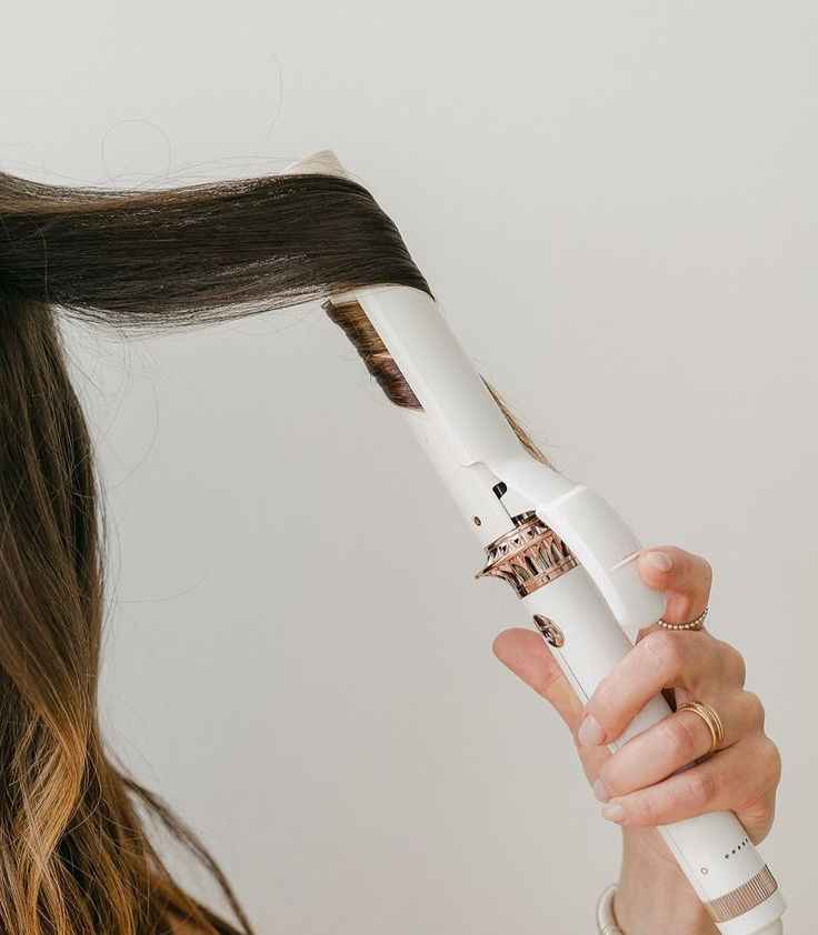 A hand uses a white curling iron on brown hair against a plain background. The curling iron has gold accents. The mood is focused.