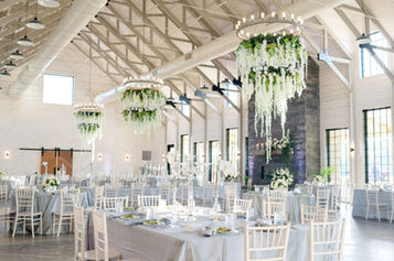 A spacious event venue with white Chiavari chairs, round tables, and grand floral chandeliers hanging from the ceiling.