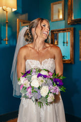 A bride in a strapless wedding dress, laughing while holding a bouquet of purple, white, and green flowers, set against a blue wall.