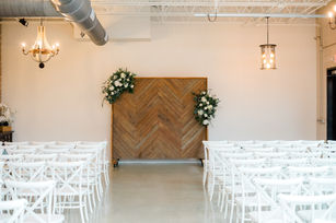 A minimalist indoor wedding ceremony space with white chairs and a wood-paneled wall adorned with florals.