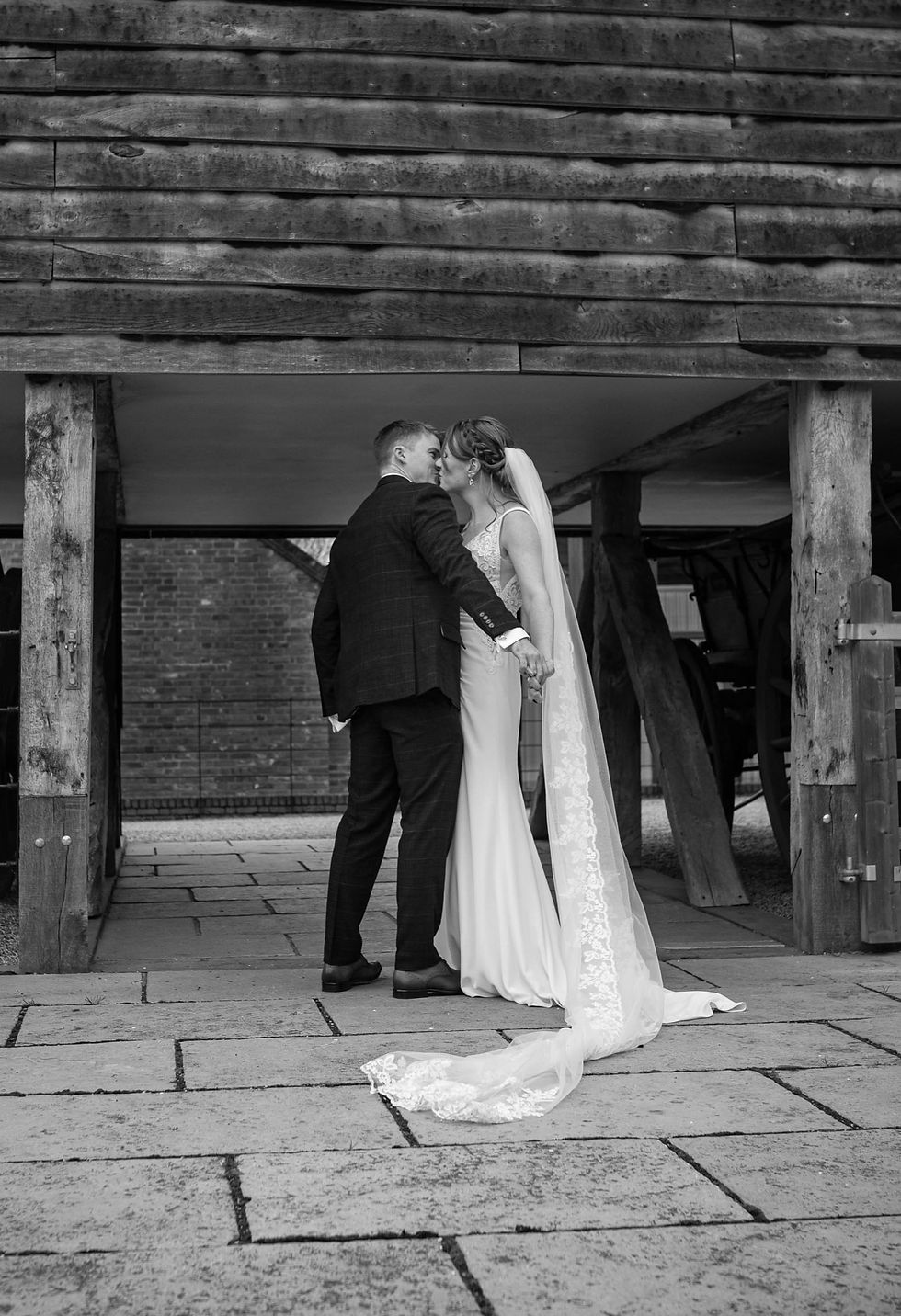 A black and white photograph of a relaxed bride and groom kissing. The bride's veil is draped across the floor in front of them.