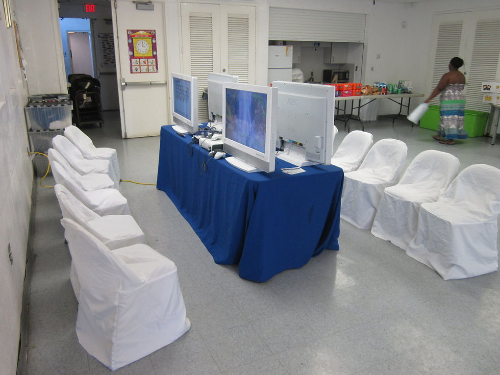 Several televisions are arranged back-to-back along a long blue-covered table, with rows of white-covered chairs on both sides in an indoor event hall. This portable console-gaming layout shows the seating and equipment arrangement for a Mobile Gaming Events Video Gaming Party.