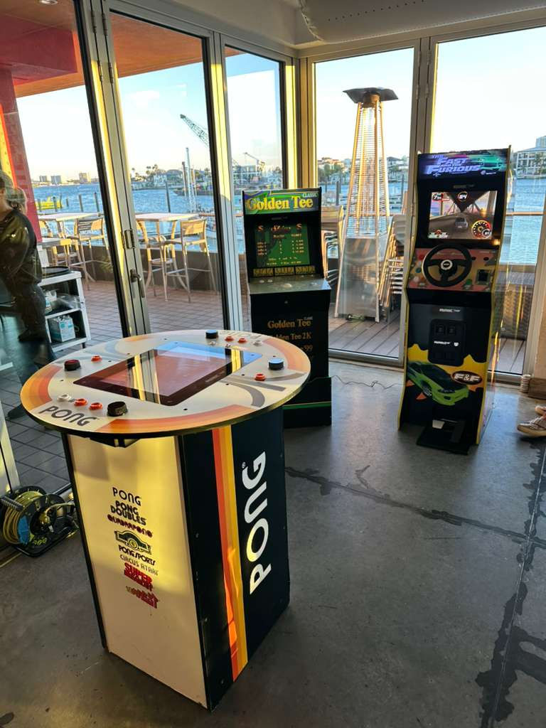 Real arcade event setup with a Pong table, Golden Tee cabinet and The Fast and the Furious racing cabinet beside floor-to-ceiling windows.