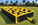 Angled view of the Maze Inflatable XL on grass, with tall black fabric walls, yellow inflatable frames, a visible entrance and a blower beside the structure.