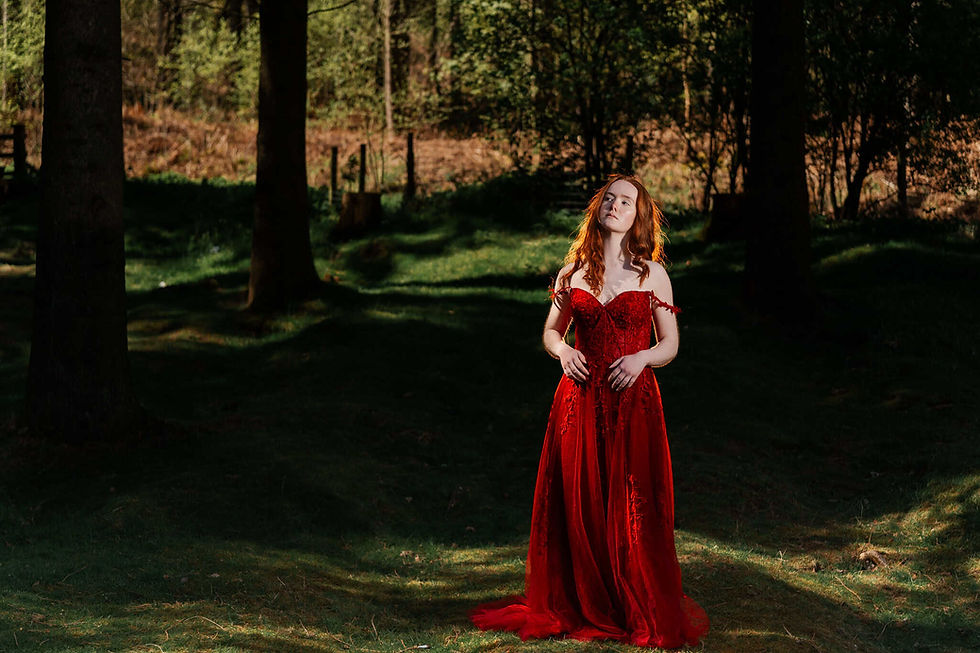 Red-haired woman in a vivid red dress stands in a sunlit forest, eyes closed, surrounded by lush greenery and shadows, feeling serene.