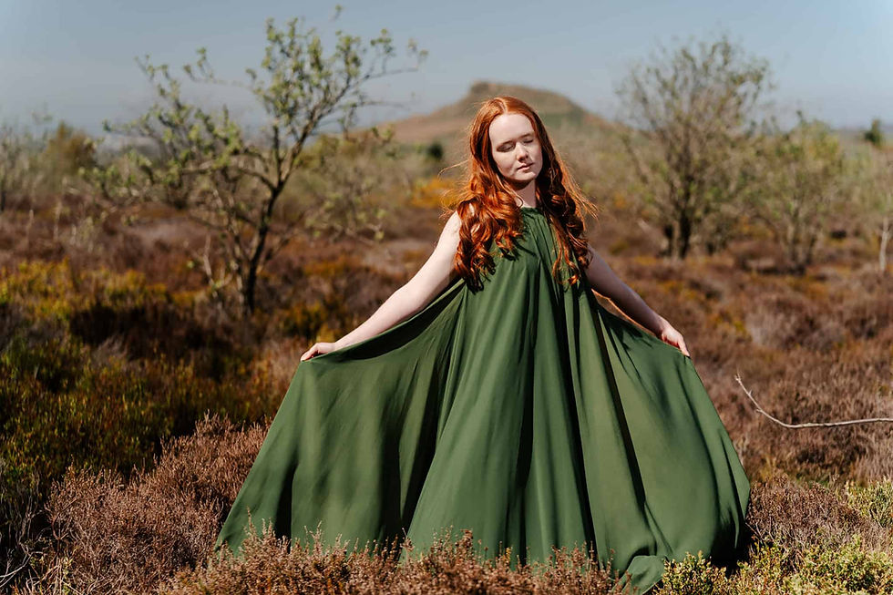 Red-haired woman in a flowing green dress stands in a grassy field, eyes closed, exuding a serene mood. Sparse trees and a hill in the background.