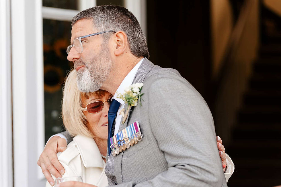 Man in gray suit with medals hugging a woman in white coat. They appear joyful in a warmly lit setting.