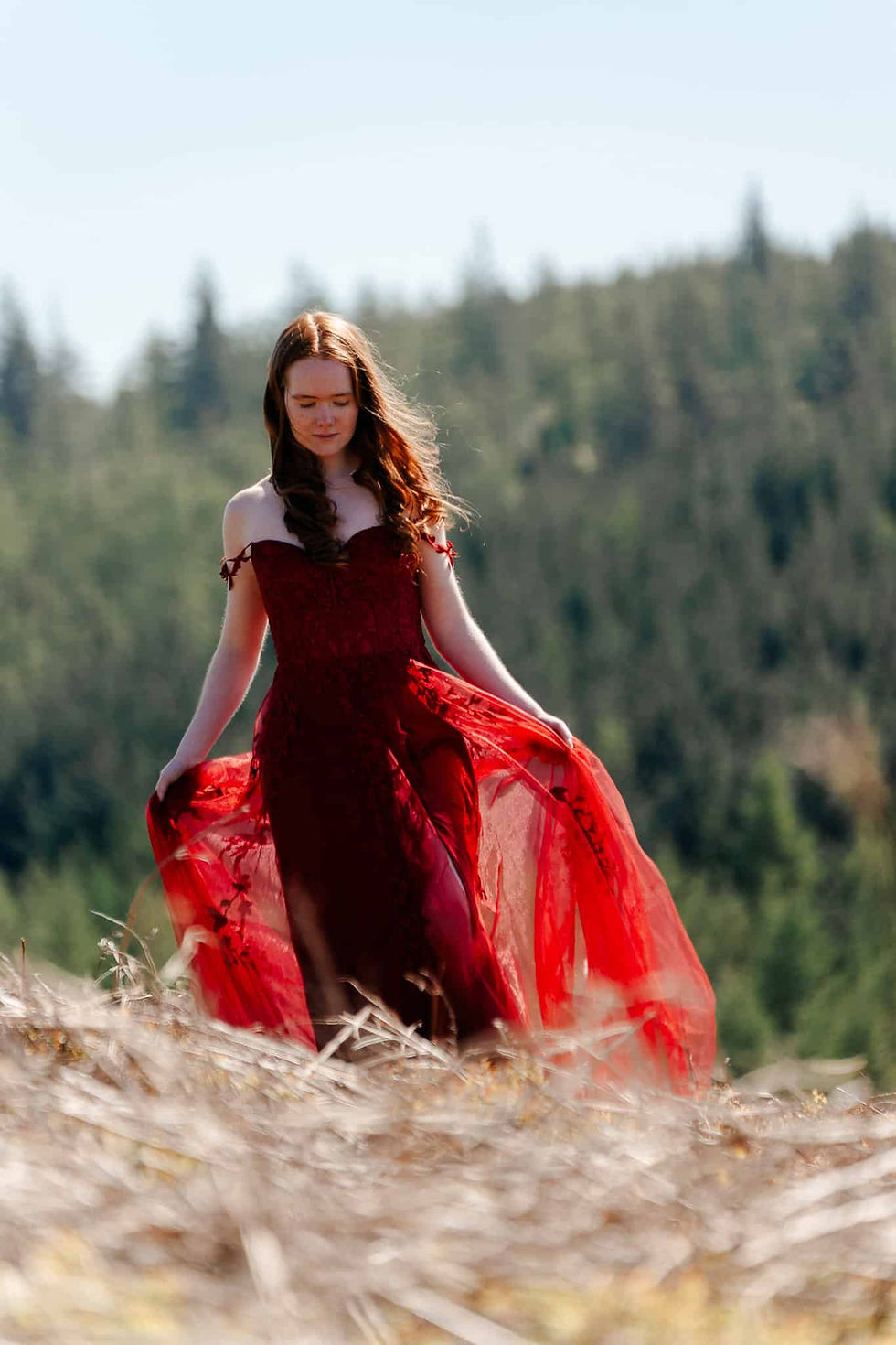 Woman in a flowing red dress stands in a field with trees in the background, appearing serene on a sunny day.