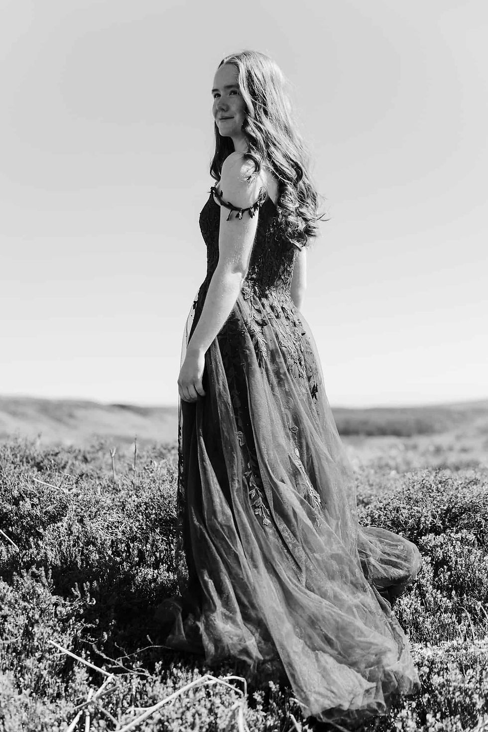Young woman in a flowing, dark gown stands in a grassy field. She looks to the side, exuding a calm, serene mood. Black and white photo.