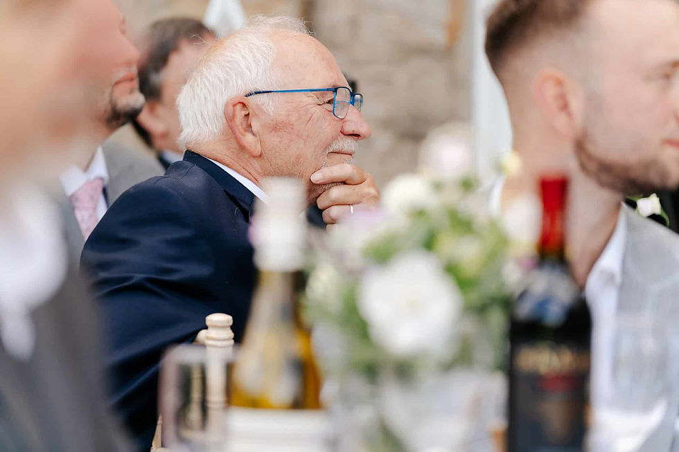 Elderly man with blue glasses in a suit, thoughtful, surrounded by people and flowers. Bottles on the table, blurred background.