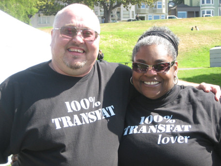 A balding white man wearing a shirt saying "100% transfat" has his arm around a black woman who wears a shirt that says "100% transfat lover"