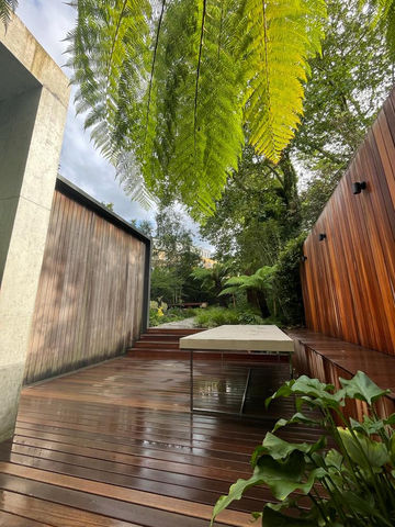 Looking through a fern leaf at a wet, yellow balau hardwood deck and fence. An outdoor table sits in front of the custom bench