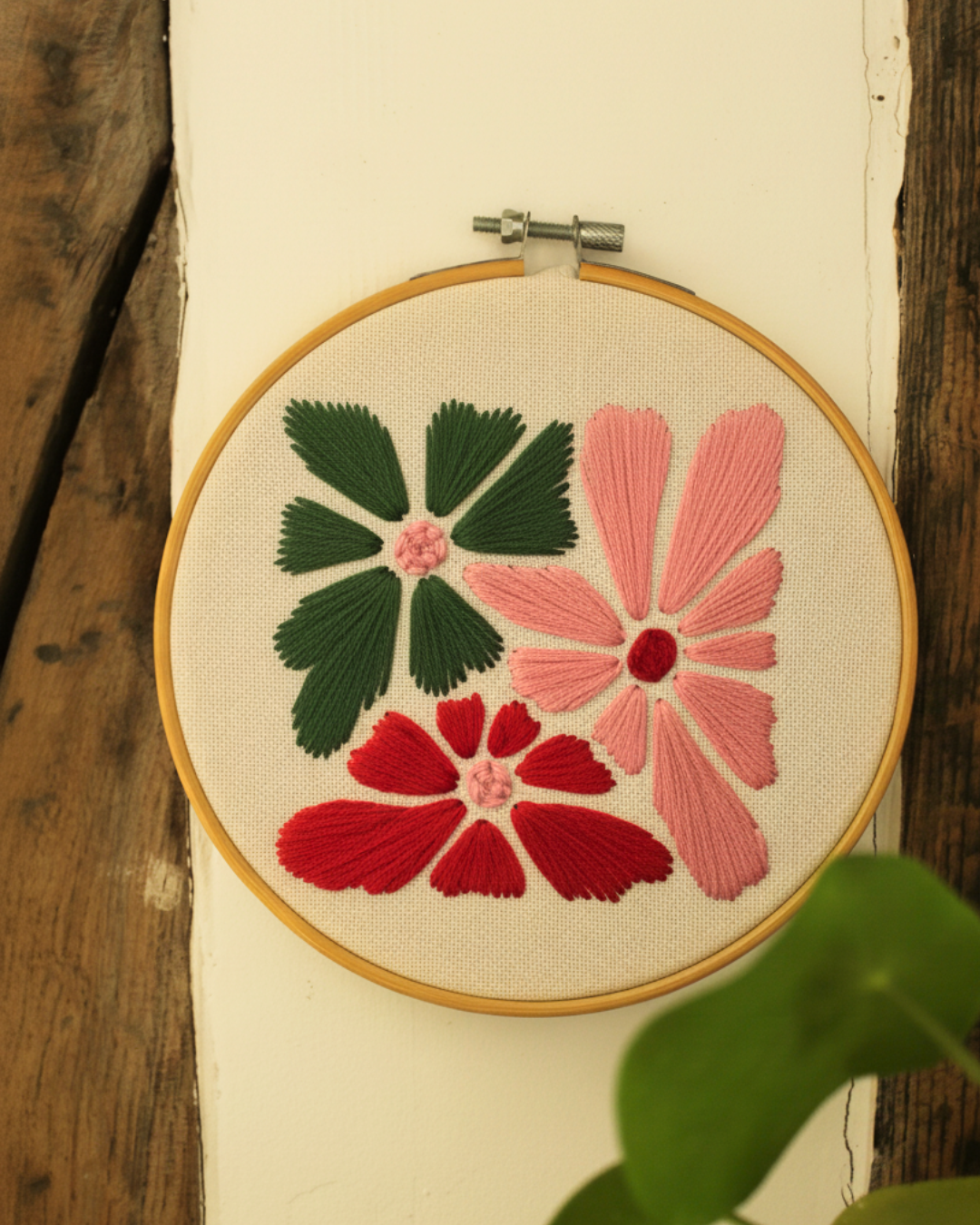 Cadre de broderie rond en bois représentant des fleurs, accroché sur un mur blanc, à côté d'une plante.