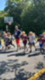 A teacher leads children running on an outdoor basketball court