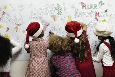 Children in festive hats color a Christmas poster reading "Merry Christmas" with drawings of Santa, reindeer, and stars in a joyful setting.
