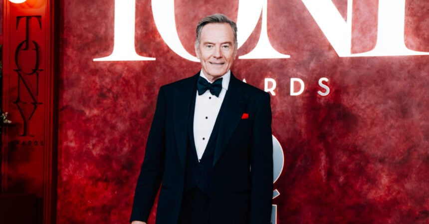 Man in tuxedo at Tony Awards red carpet, standing before a red backdrop with "TONY" in large white letters. Elegant and formal setting.