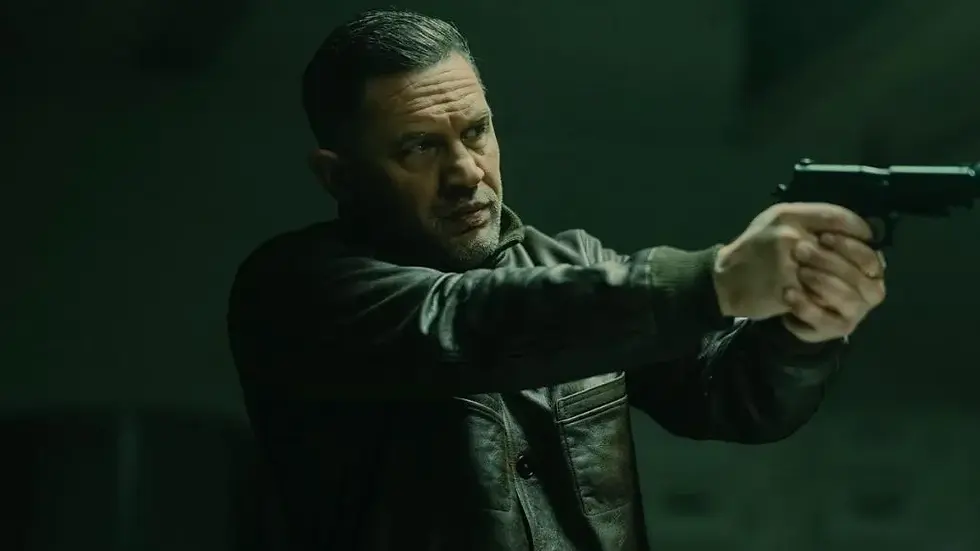 Man in dark jacket aiming a pistol in a dim, green-lit room, appearing tense and focused. No visible text.