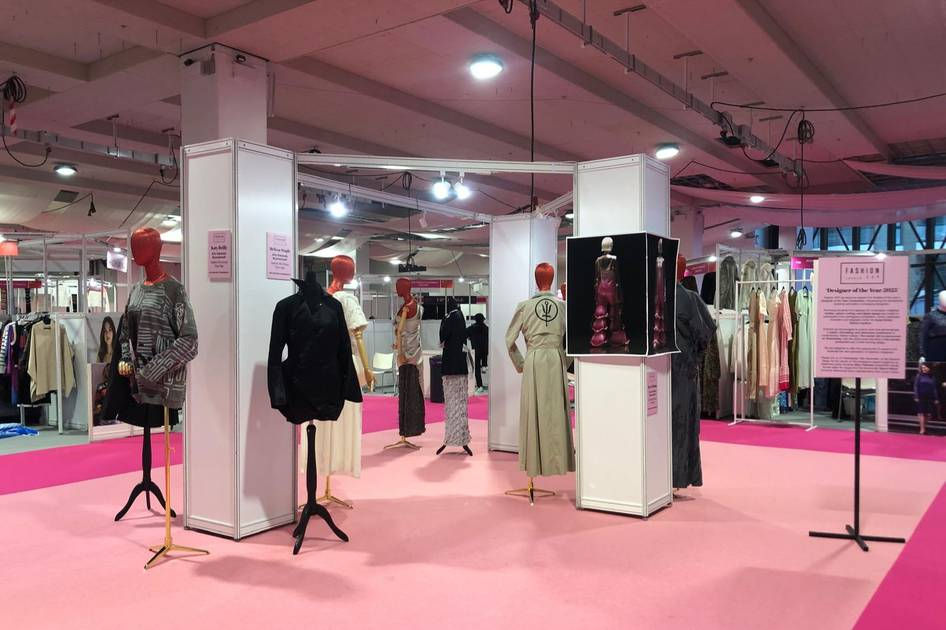 Mannequins showcase diverse clothing on a pink floor in a fashion exhibit. Bright lighting enhances the colorful outfits and informational signs.
