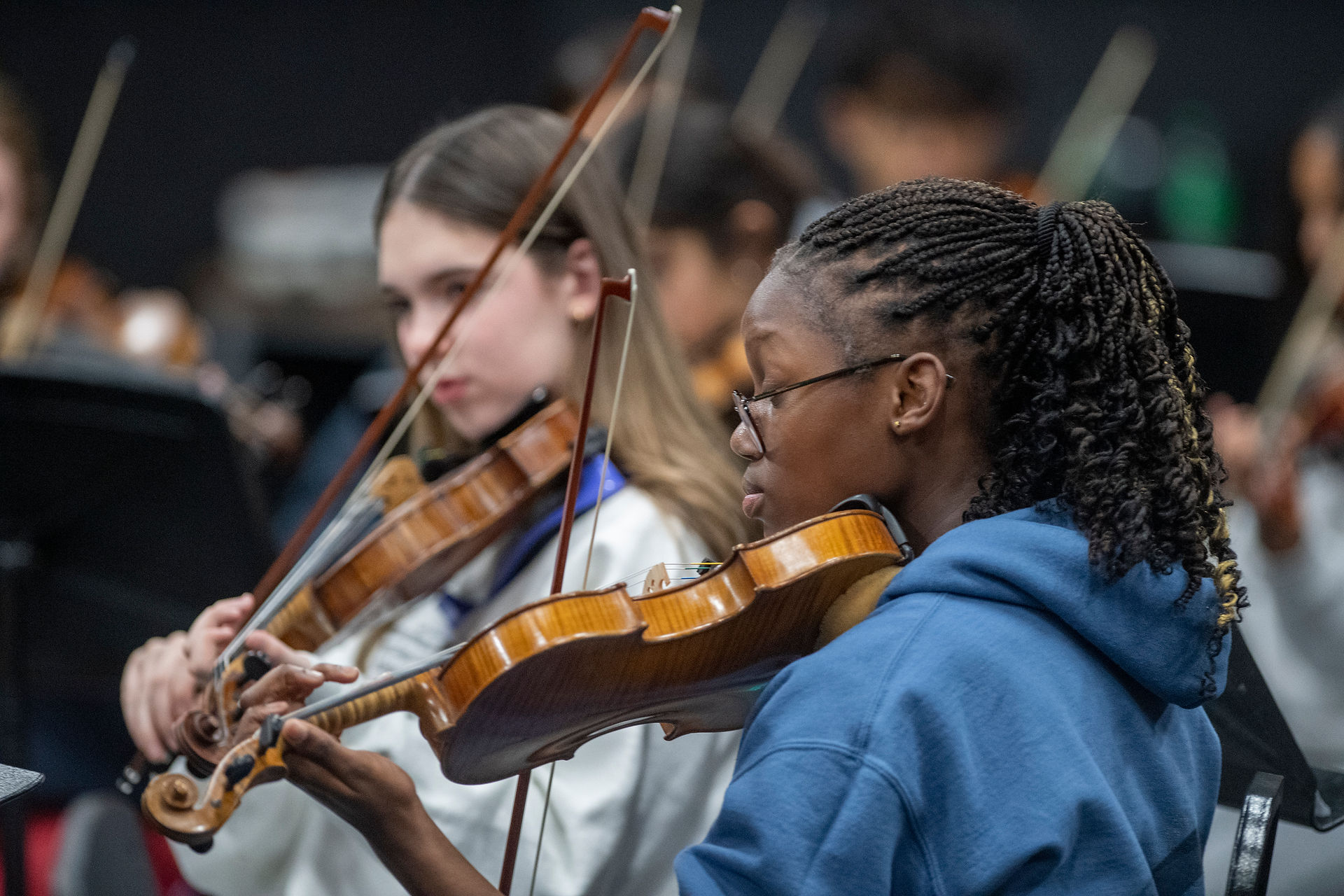 Close-up of two young violinists rehearsing; a student in a blue hoodie and glasses plays in the foreground while another violinist is softly blurred behind
