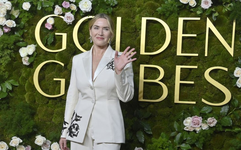 “Actor in a tailored white suit waving on a Golden Globes red carpet, standing in front of a lush green floral wall with the awards logo.”
