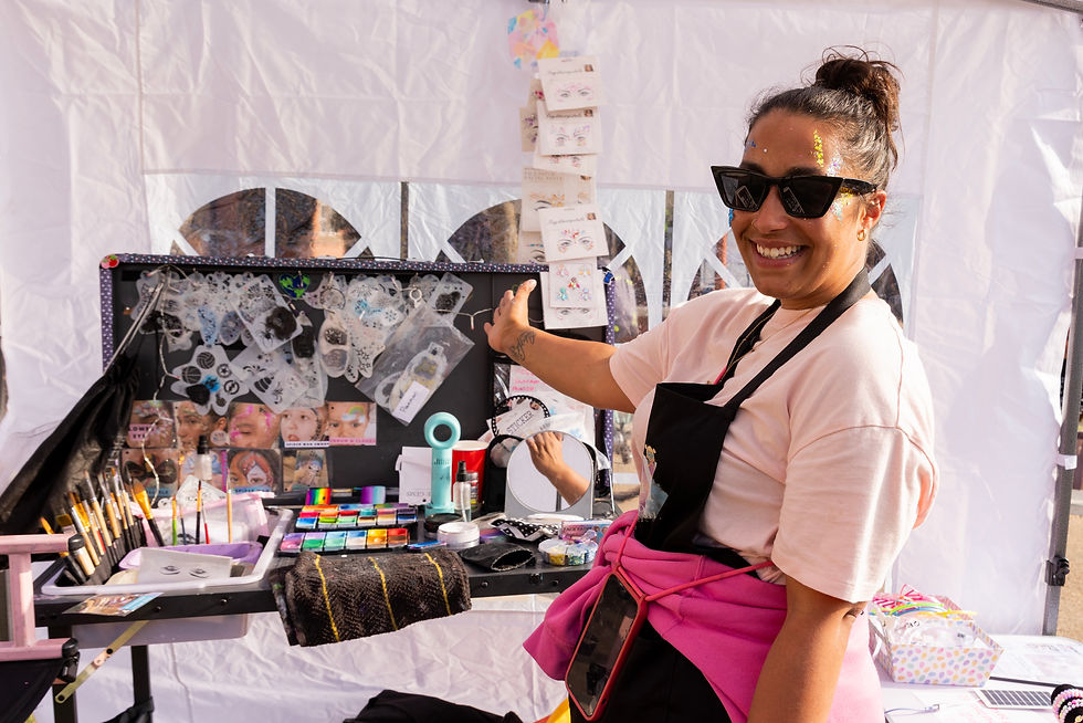 Smiling woman in sunglasses and apron points to a face painting setup with colors and stencils in a sunny outdoor tent at Brew Beat Leicester festival.