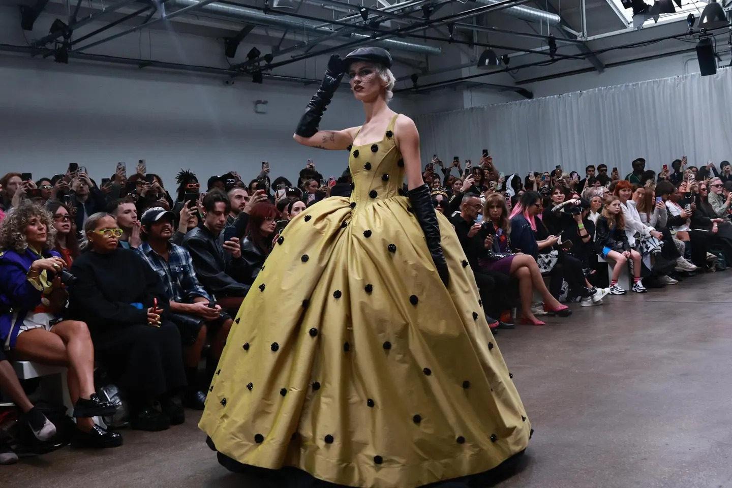 A model in a dramatic yellow ball gown with black polka dots and long black gloves walking past a front row of guests all filming the London Fashion Week show.   Alt text 6: A model in a black velvet gown with bold white shoulder detail and a large flower motif walking a floral-lined runway during an elegant fashion presentation.
