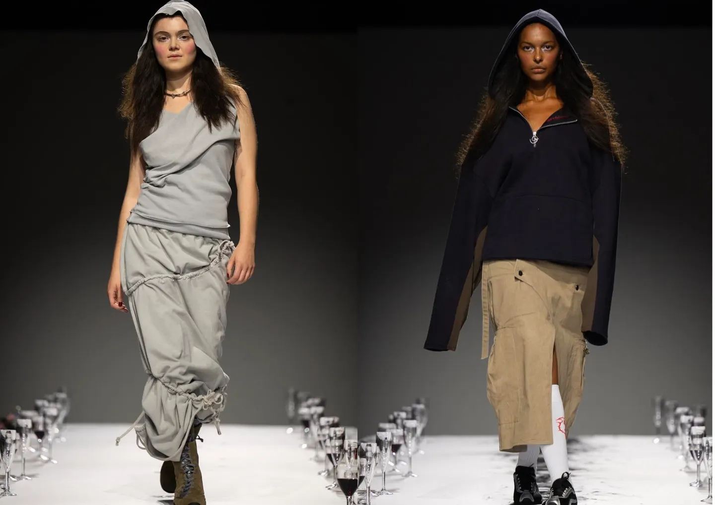Two models on a catwalk lined with wine glasses, wearing deconstructed hooded streetwear – one in a draped grey skirt set, the other in an oversized black hoodie with tan cargo skirt.