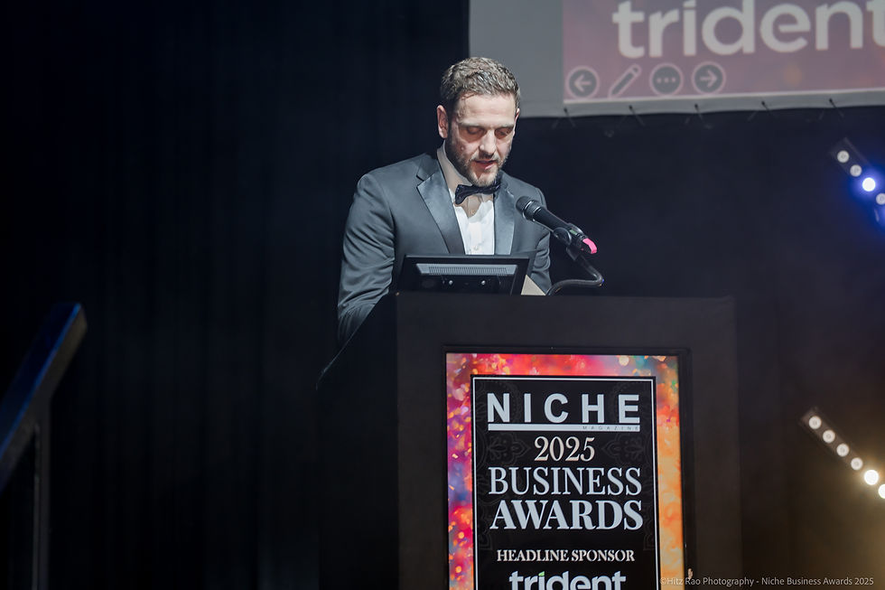 Adam Burrage
in a tuxedo speaks at a podium with "NICHE 2025 BUSINESS AWARDS" text. Dimly lit stage, dark background, award event mood.