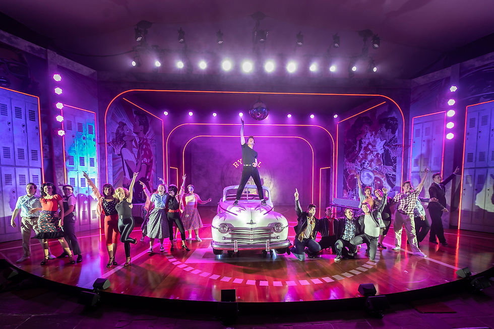 Grease performers in vibrant 50s attire dance energetically on stage. One stands on a vintage car. Bright lights and colorful set evoke excitement at Kilworth House Theatre