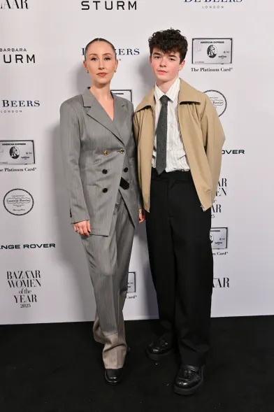 Two guests on the Harper’s Bazaar Women of the Year 2025 red carpet; one in an oversized grey double-breasted suit and the other in a light beige jacket over a shirt and tie, standing in front of the branded backdrop
