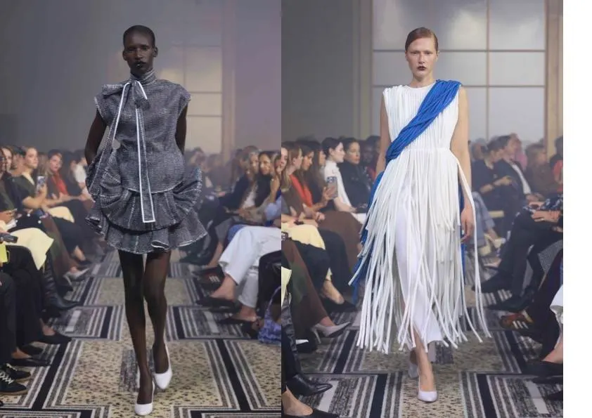 Two models walking a packed runway show, one in a sculptural grey ruffled mini dress, the other in a white dress with long shredded fringe and a blue sash.