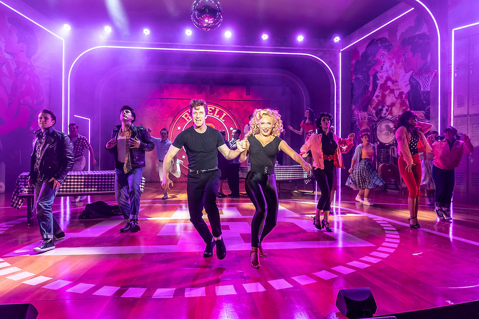 Grease dancers perform energetically on stage with neon lights and a disco ball. The backdrop features vibrant murals and the word "Rydell" at Kilworth House Theatre.